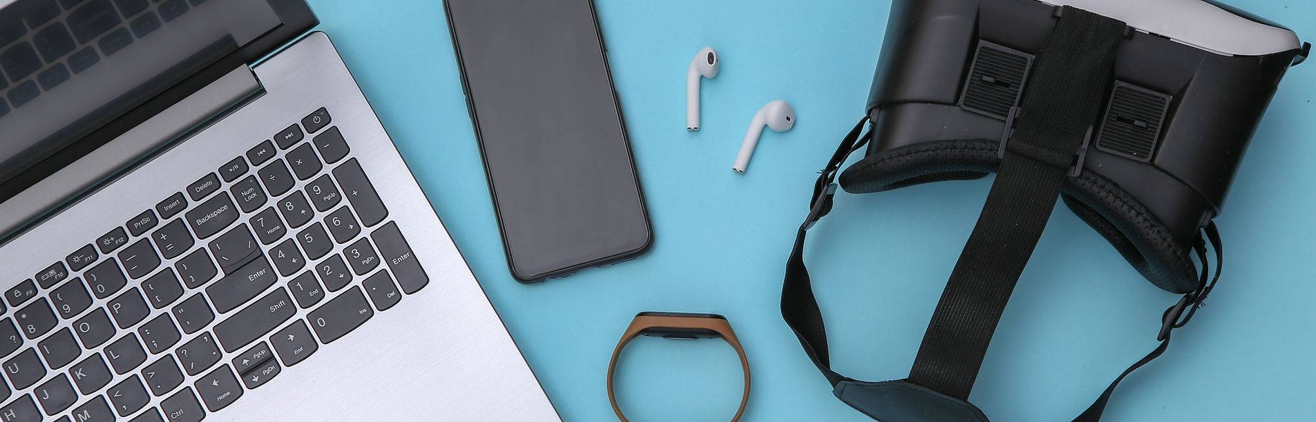 A laptop, smartphone, wireless earbuds, fitness band and virtual reality headset on a blue surface