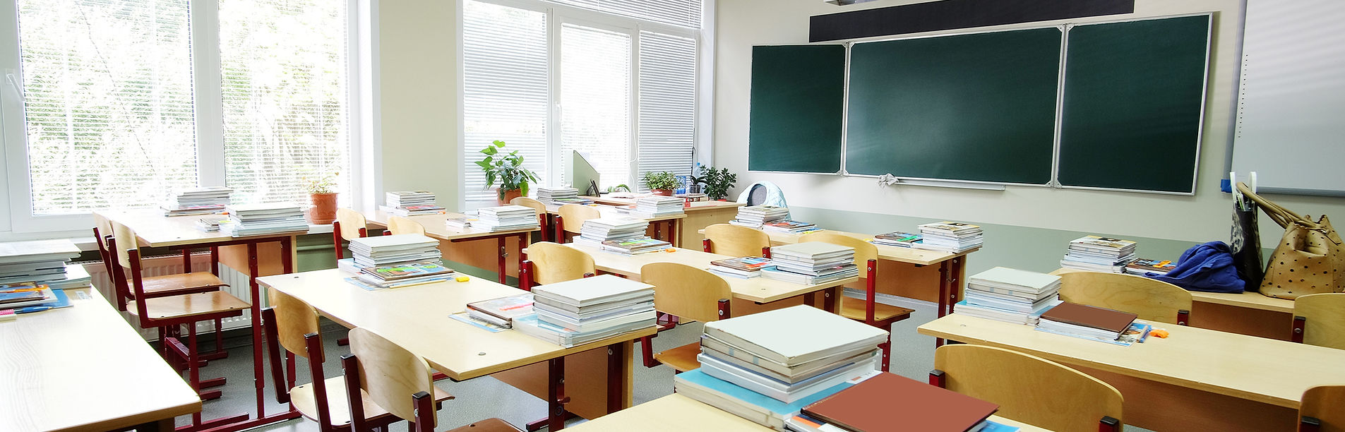 An empty classroom with rows of desks stacked with books and a chalkboard at the front