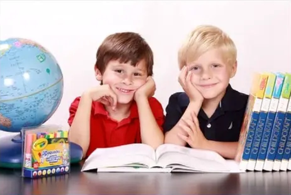 Two young children reading a book at a table beside a globe and workbooks