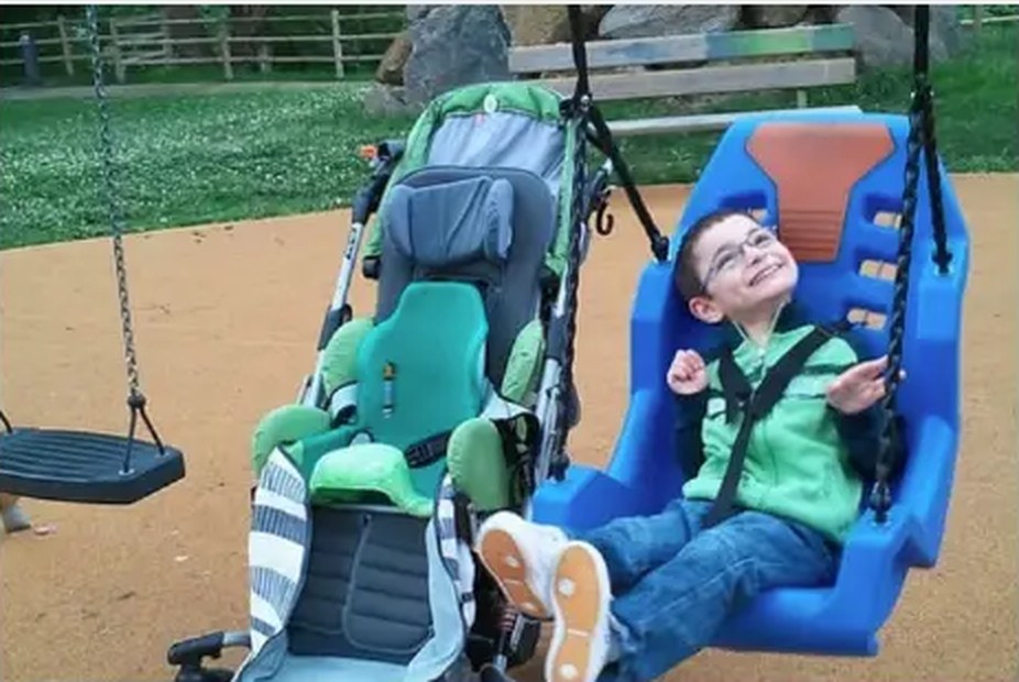 A smiling child seated in an adaptive playground swing next to an adaptive stroller